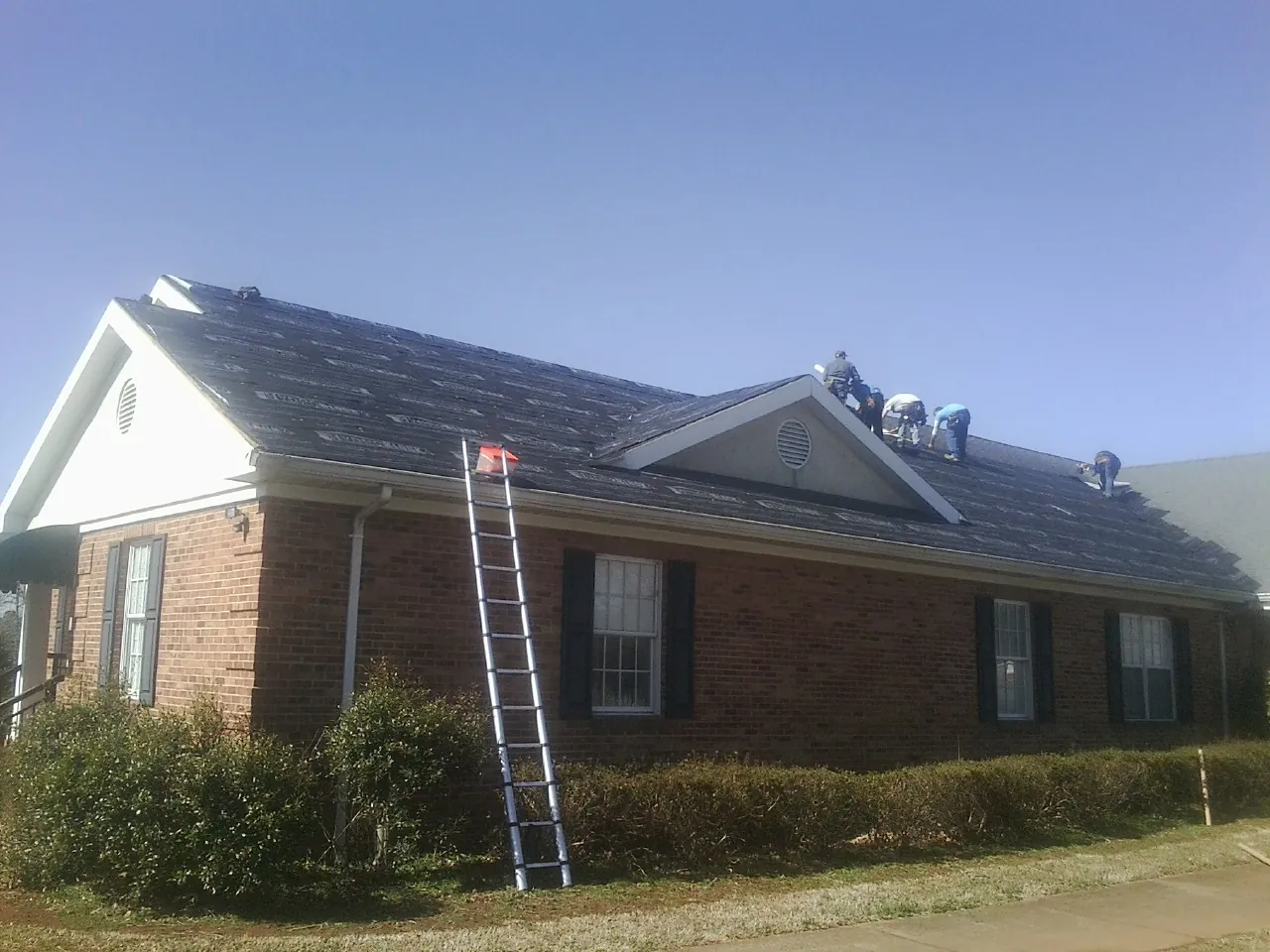 Crew tearing shingles off a brick ranch roof with a ladder set — Emergency Roof Repair work by our crew in Grand Blanc