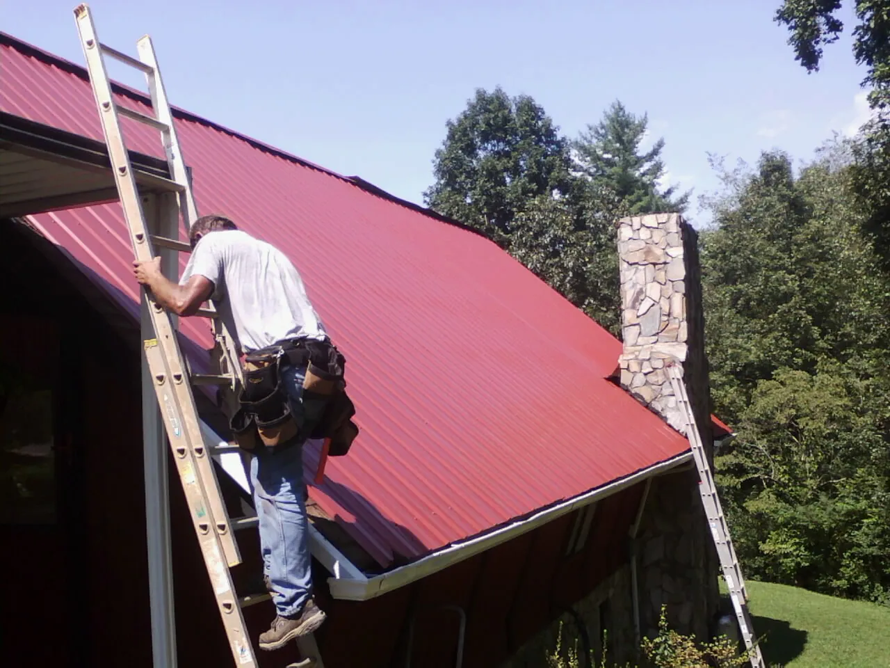 Roofer on a ladder beside a red metal roof and stone chimney — Soffit & Fascia Repair by Roofing Brothers in Grand Blanc